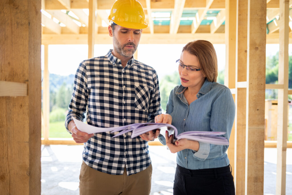 Construction contractor wearing a yellow hard hat reviewing building plans with a woman at a residential construction site.